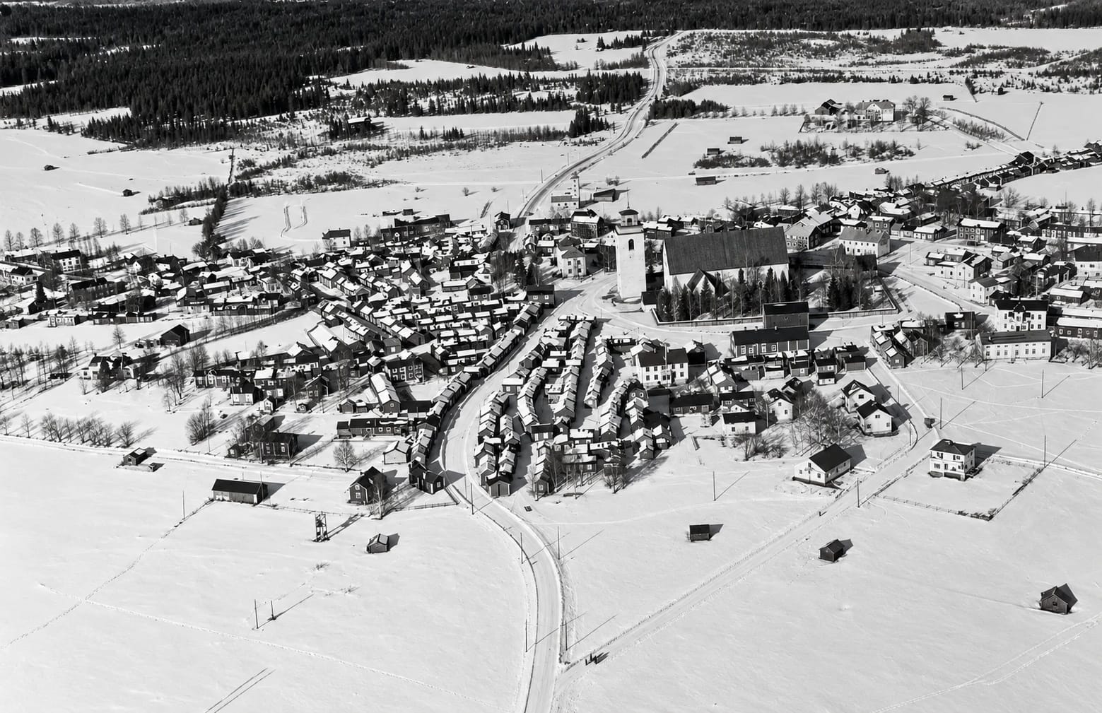 Flygfoto i svartvitt över Gammelstads kyrkstad en vinterdag, med kyrkan, kyrkstugorna, vägar och snötäckta åkrar.
